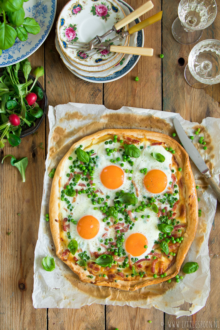 Verwarm de oven voor op 190 graden. Blancheer de erwtjes kort en spoel af onder de koude kraan. Leg het quichedeeg op een bakblik. Besmeer het deeg met de crème fraîche. Verdeel hierover de erwtjes en spekjes. Zet de flammkuchen 20 minuten in de oven. Haal eruit, breek de eitjes erop en breng op smaak met peper en zout. Zet nog eens 15 minuten in de oven Garneer met basilicum en snijd in punten.